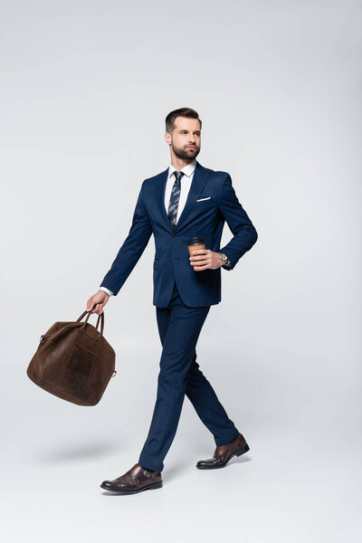 economist in blue suit walking with coffee to go and leather briefcase on grey