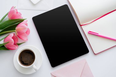 Pembe lalelerin yanındaki kahve fincanının üst görüntüsü ve boş ekranlı dijital tablet ve beyaz üzerine not defteri. 