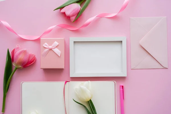 镜框附近的郁金香 空白笔记本 信封和粉红礼品盒的顶部视图 — 图库照片