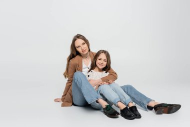 Gri arka planda oturan modaya uygun giyinmiş anne ve çocuğun tam boy görüntüsü