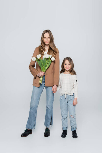 full length view of stylish woman with tulips holding hands with daughter on grey background