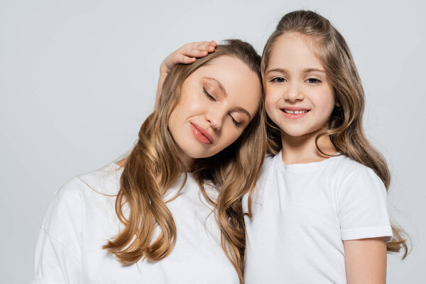 cheerful girl looking at camera near mother with closed eyes isolated on grey