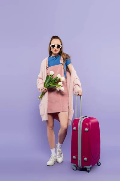 全身上下都是身穿粉红连衣裙 戴着太阳镜的女人 站在紫色行李旁边 带着鲜花 — 图库照片