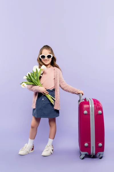 全身上下都是快乐的女孩 戴着太阳镜 带着鲜花站在紫色的粉红行李旁边 — 图库照片