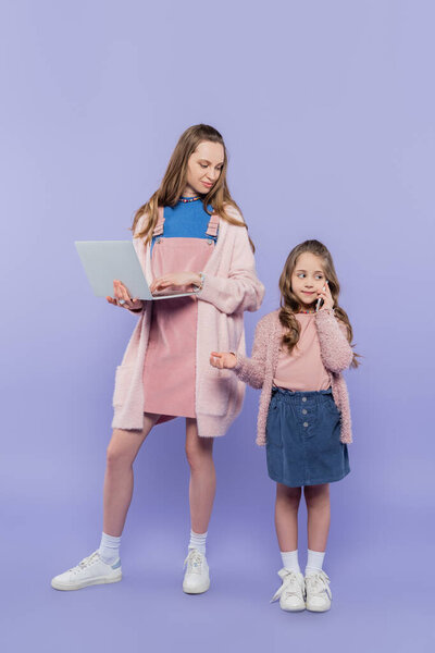 Мать с ноутбуком в полный рост смотрит на ребенка, разговаривающего по смартфону на фиолетовом 