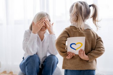 At kuyruklu bir kız elinde mavi ve sarı bir kalple mutlu bir annenin yanında elleri ve yüzü belirsiz bir şekilde kart tutuyor.