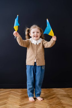full length view of joyful patriotic child holding ukrainian flags near black wall clipart