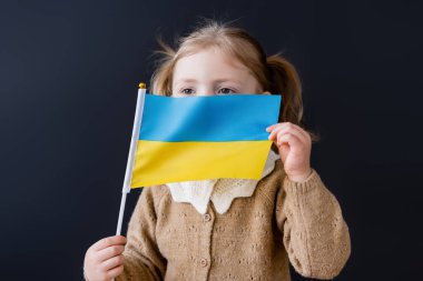 Siyah üzerine izole edilmiş küçük Ukrayna bayrağıyla yüzünü gizleyen küçük bir kız.