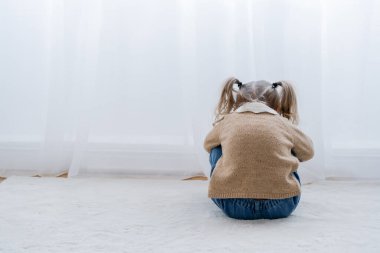 Beyaz perdenin yanında yerde oturan üzgün çocuğun arka görünüşü