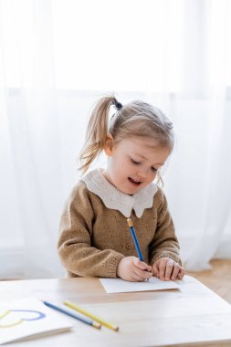Neşeli çocuk, masasında mavi ve sarı bir kalp olan kartın yanında resim çiziyor.
