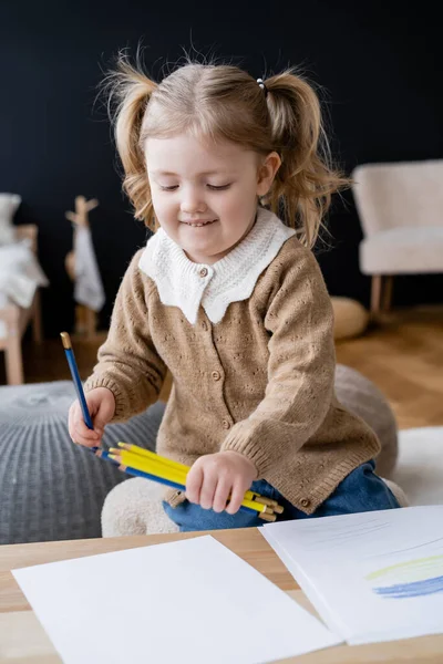 笑容可亲的女孩 马尾辫上挂着蓝色和黄色的铅笔 接近模糊的桌子 — 图库照片