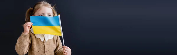 patriotic child obscuring face with small ukrainian flag isolated on black with copy space, banner