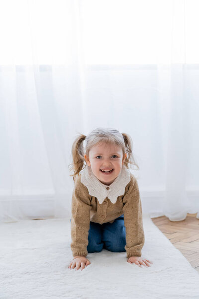 cheerful girl with ponytails crawling on floor at home and looking at camera