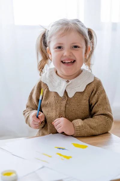 一个快乐的女孩在纸旁边拿着画笔 涂满了蓝色和黄色的东西 — 图库照片