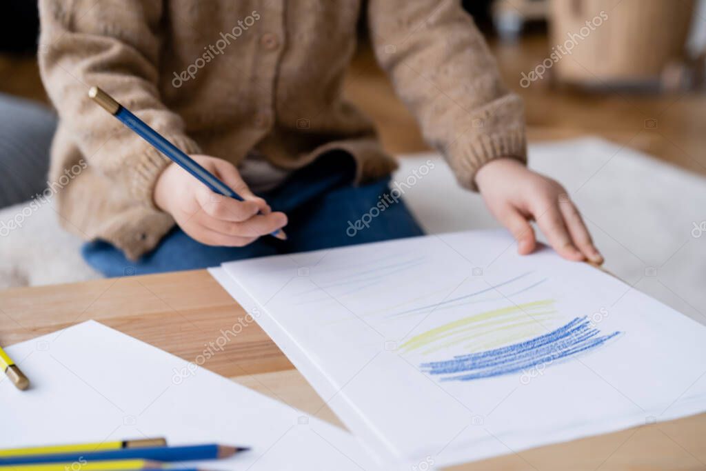 cropped view of a blurred child holding color pencil near papers with blue and yellow strokes