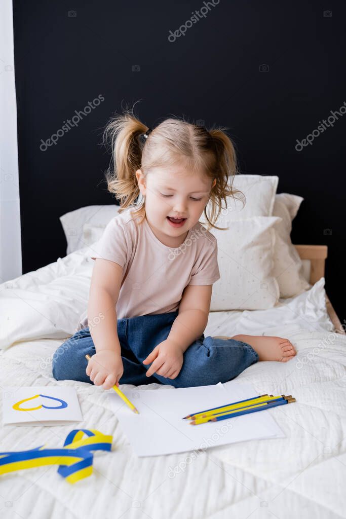 happy girl drawing with color pencil near ribbon and card with blue and yellow heart 