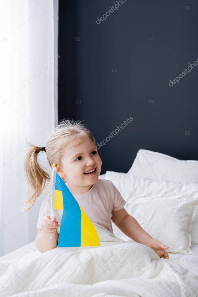 cheerful girl holding small ukrainian flag while sitting on bed