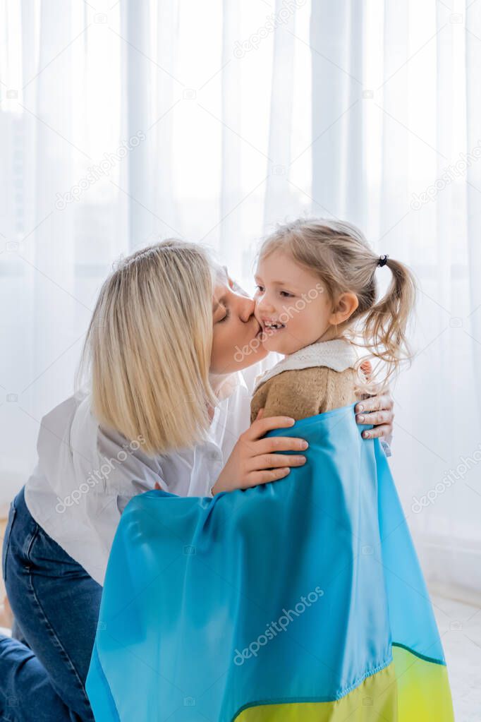 blonde woman kissing cheerful daughter covered with ukrainian flag