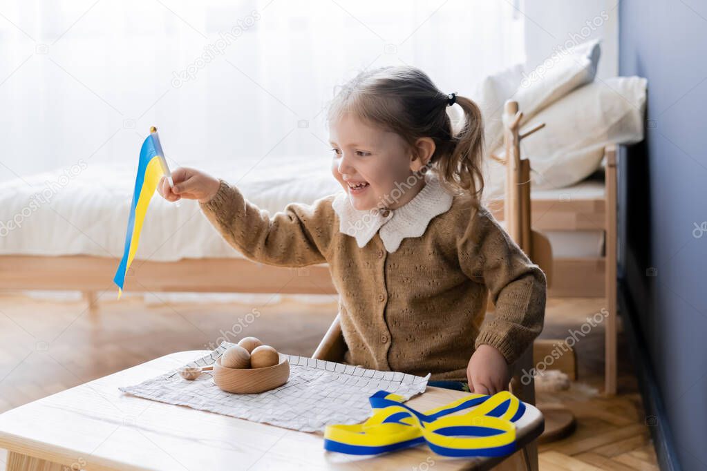 happy girl holding ukrainian flag near blue and yellow ribbon and wooden toys on desk