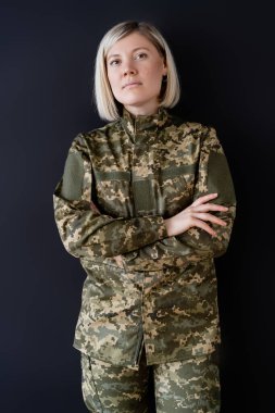 Sarışın asker kadın kollarını kavuşturup siyah kameraya bakıyor.