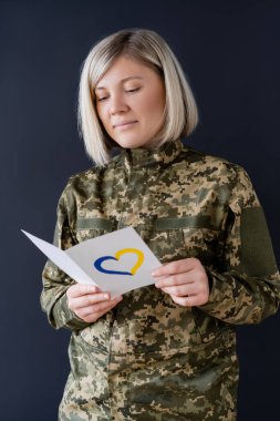 woman in military uniform reading postcard with blue and yellow heart isolated on black clipart