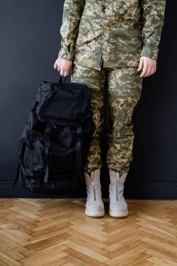 Kara duvarın yanında siyah sırt çantalı asker kadın görüntüsü.
