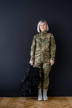 full length of woman in military uniform standing with black backpack near dark wall clipart