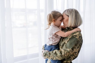 Evde askeri üniformayla annesini kucaklayan küçük kızın yan görünüşü.