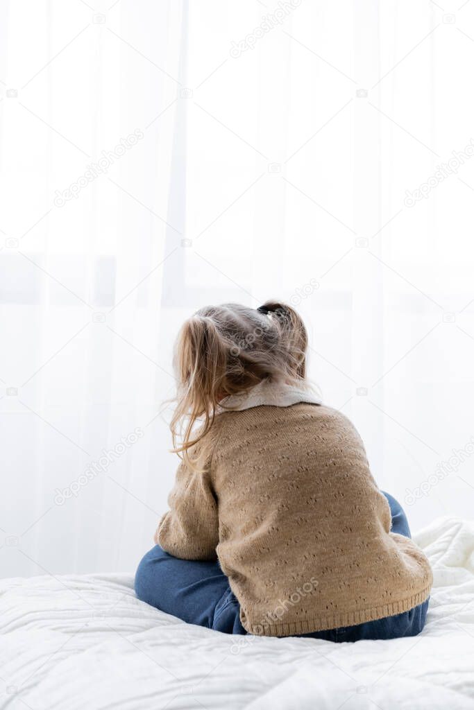 back view of little girl with ponytails sitting alone on bed at home