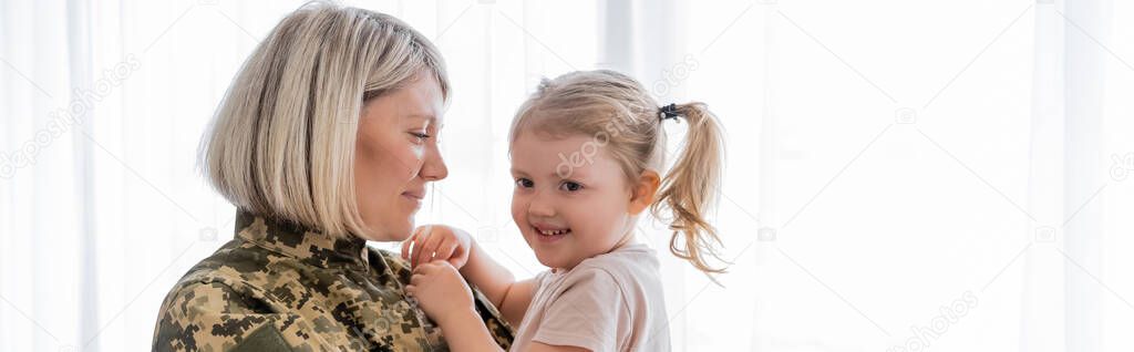 positive military woman holding happy daughter at home, banner