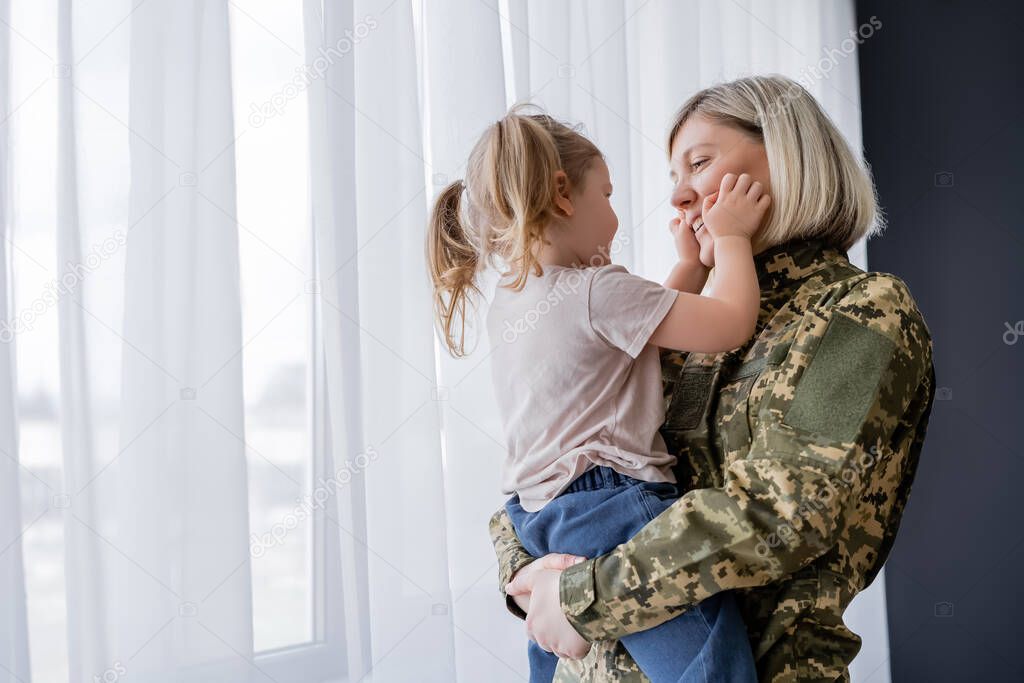 little girl with ponytails touching face of smiling mom in military uniform near window