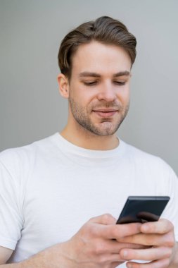 Beyaz tişörtlü genç adam gri üzerinde izole akıllı telefon kullanıyor.