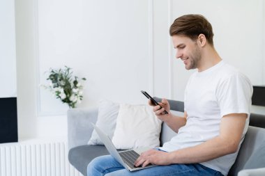 Gülümseyen adamın yan görüntüsü akıllı telefon ve dizüstü bilgisayarla evdeki kanepede oturuyor.