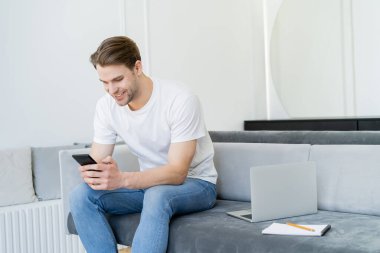 Gülümseyen adam dizüstü bilgisayarın yanında oturuyor ve akıllı telefondan mesaj yolluyor.