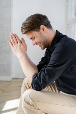 side view of man with praying hands smiling with closed eyes at home clipart