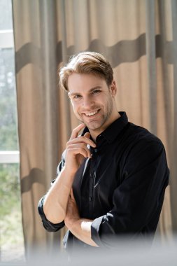 Siyah tişörtlü gülümseyen adam, yüzün yanında el ele tutuşuyor ve bej perdenin yanındaki kameraya bakıyor.