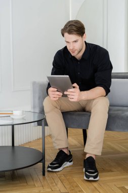 Dijital tabletli şık bir adamın oturma odasındaki sehpanın yanında oturması.
