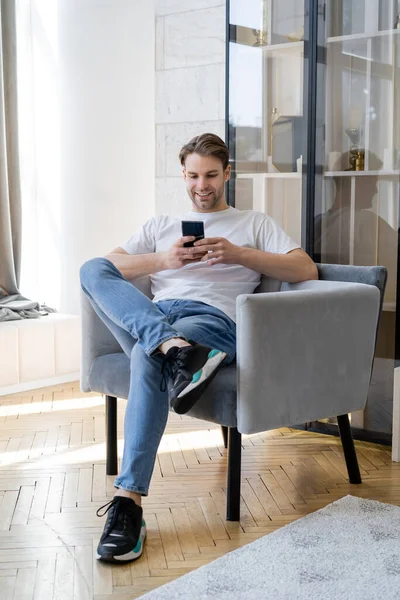 Smiling Man Jeans Sitting Armchair Crossed Legs Chatting Smartphone — Stock Photo, Image