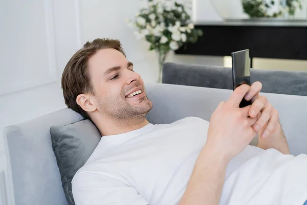 Potěšený Mladý Muž Odpočívá Gauči Chatuje Smartphonu — Stock fotografie