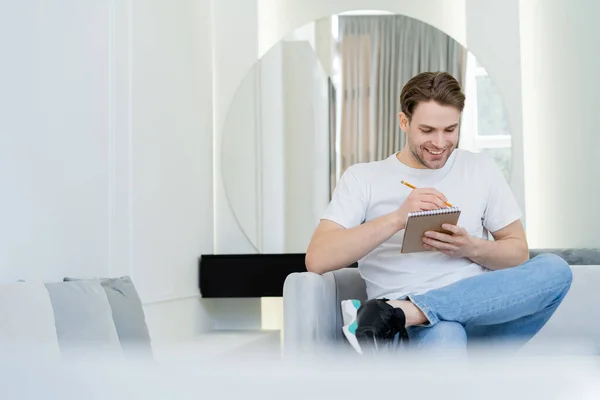 Cheerful Man Writing Notebook While Sitting Living Room Blurred Foreground — Stock Photo, Image