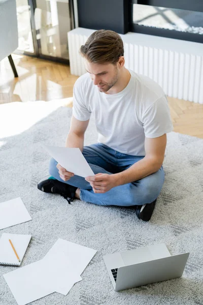 ノートパソコンの近くの床に座って文書を見ている男の高い角度の眺め — ストック写真