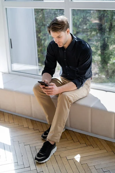 Siyah Gömlekli Pantolonlu Bir Adam Pencere Eşiğinde Oturuyor Akıllı Telefondan — Stok fotoğraf