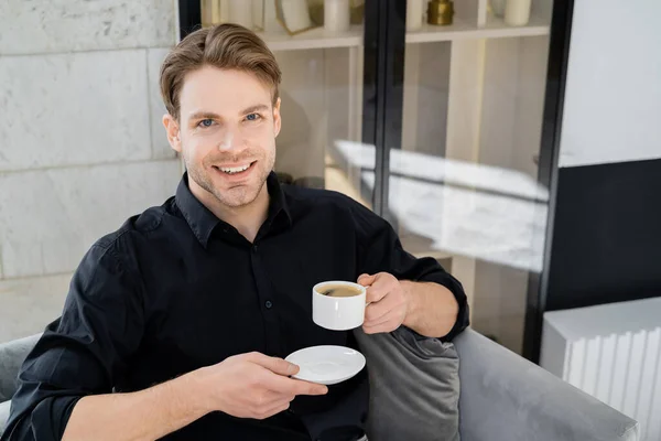 Kahve Fincanı Çay Tabağı Taşıyan Neşeli Adam Evde Otururken Kameraya — Stok fotoğraf