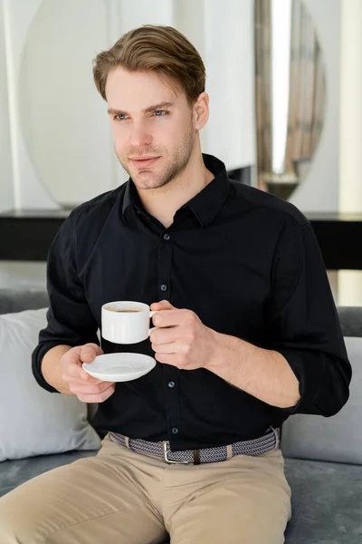 Muž Černé Košili Sedí Šálkem Kávy Podšálkem Gauči Obývacím Pokoji — Stock fotografie