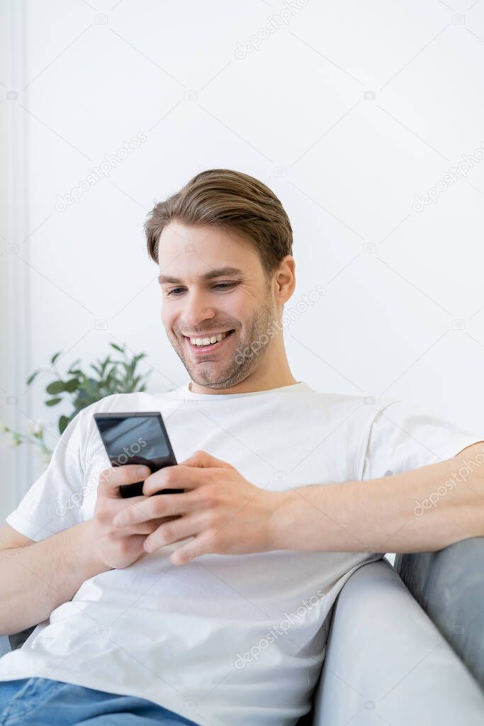 cheerful young man in white t-shirt messaging on cellphone while sitting at home