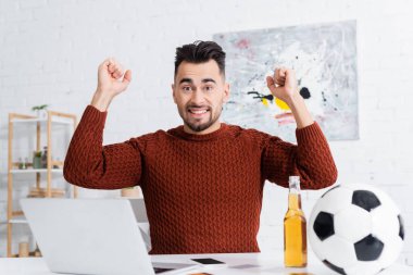 Heyecanlı bahisçi dizüstü bilgisayar, bira ve futbol topu yakınlarında başarı jesti sergiliyor
