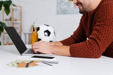 cropped view of smiling bookmaker typing on laptop near credit cards and soccer ball