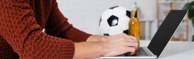 partial view of bookmaker typing on laptop near soccer ball and beer, banner