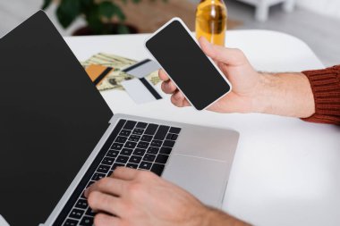 Akıllı telefonlu bahisçinin kısmi görüntüsü ve bulanık kredi kartlarının yanındaki boş ekranlı dizüstü bilgisayar