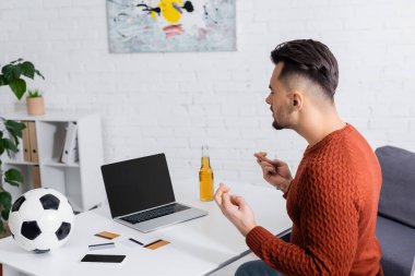 gambler with crossed fingers near laptop with blank screen, credit cards and soccer ball